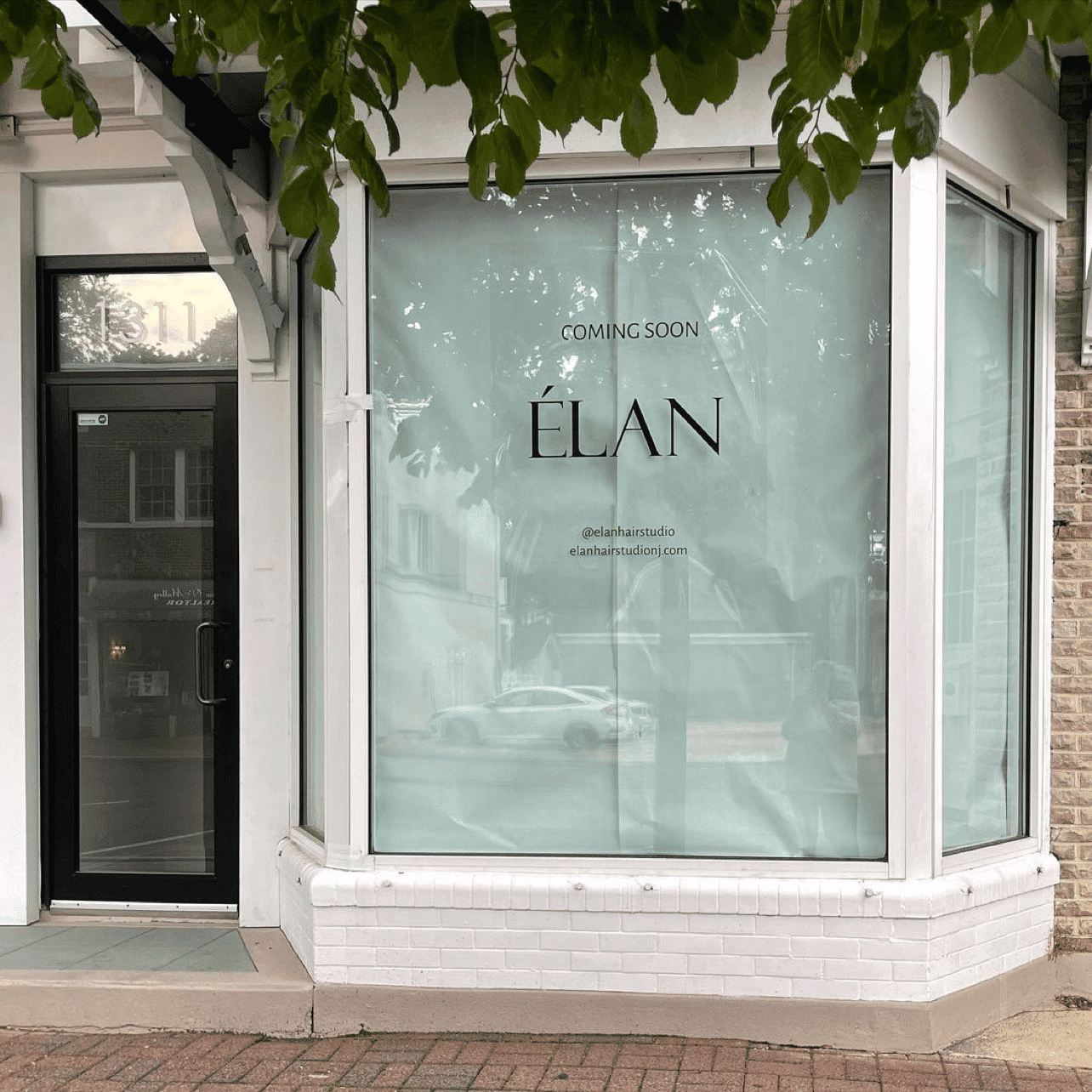 New store "Élan" coming soon, sign on a storefront window with leafy green branches above.