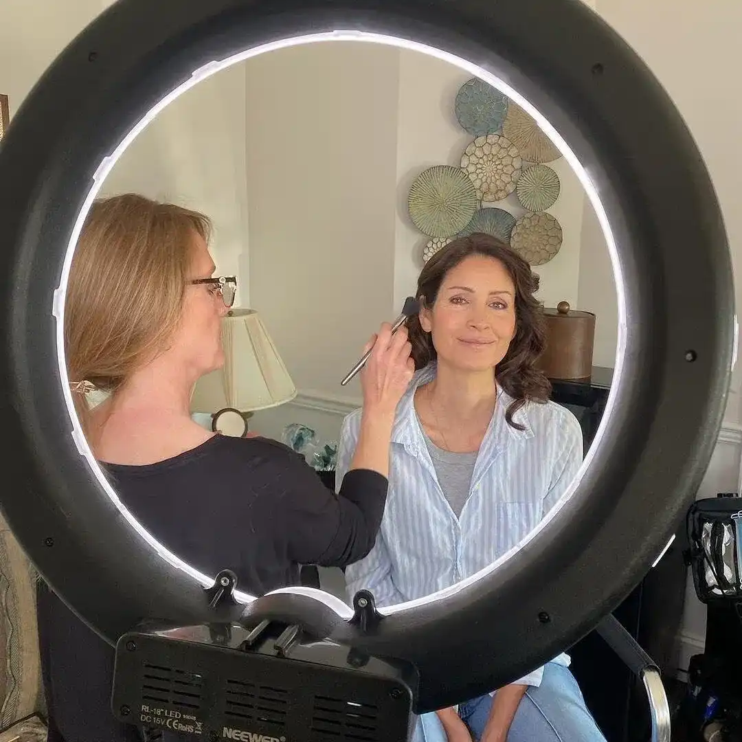 Makeup artist applies makeup on a smiling woman in front of a ring light.