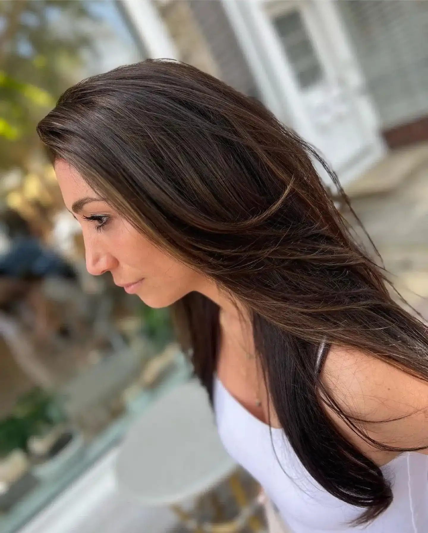 Woman with long brown hair in profile, wearing a white top, outdoors.